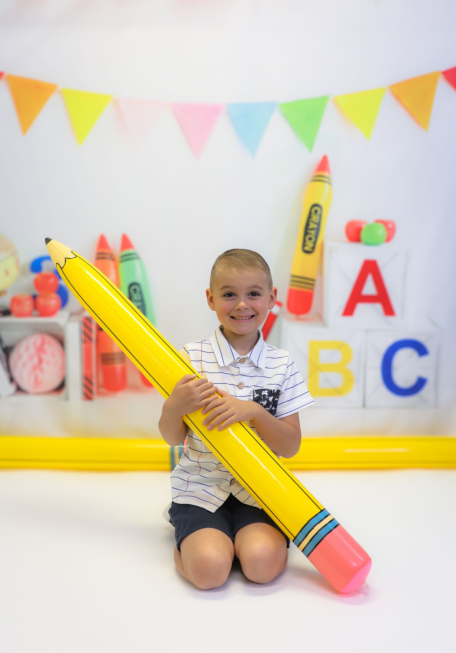A child kneels on the floor holding a large yellow pencil prop against a white wall with colorful ABC decorations.