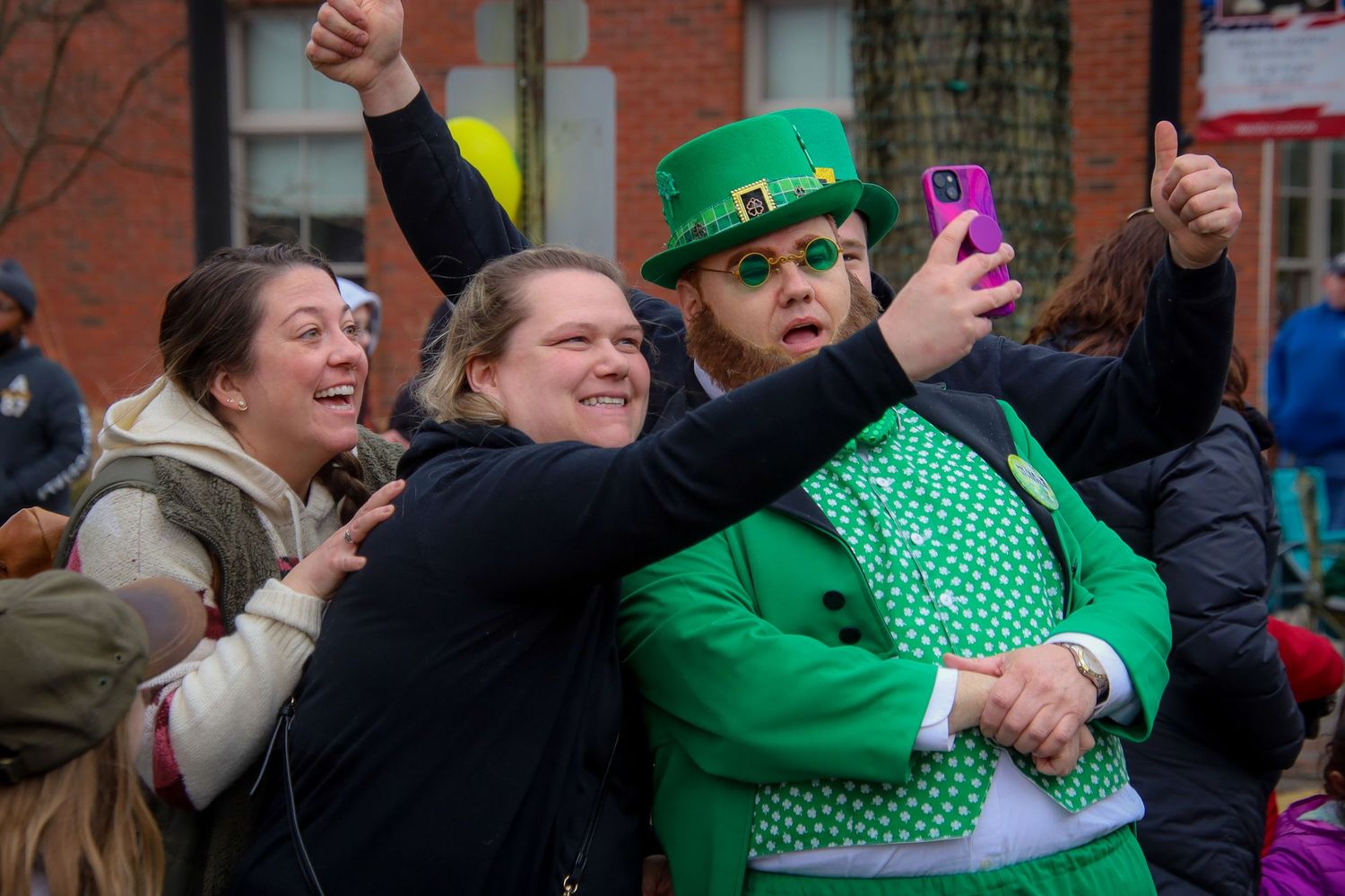 Festive crowd celebrates at St Patrick's Day parade wearing bright green attire and taking photos with smartphones.
