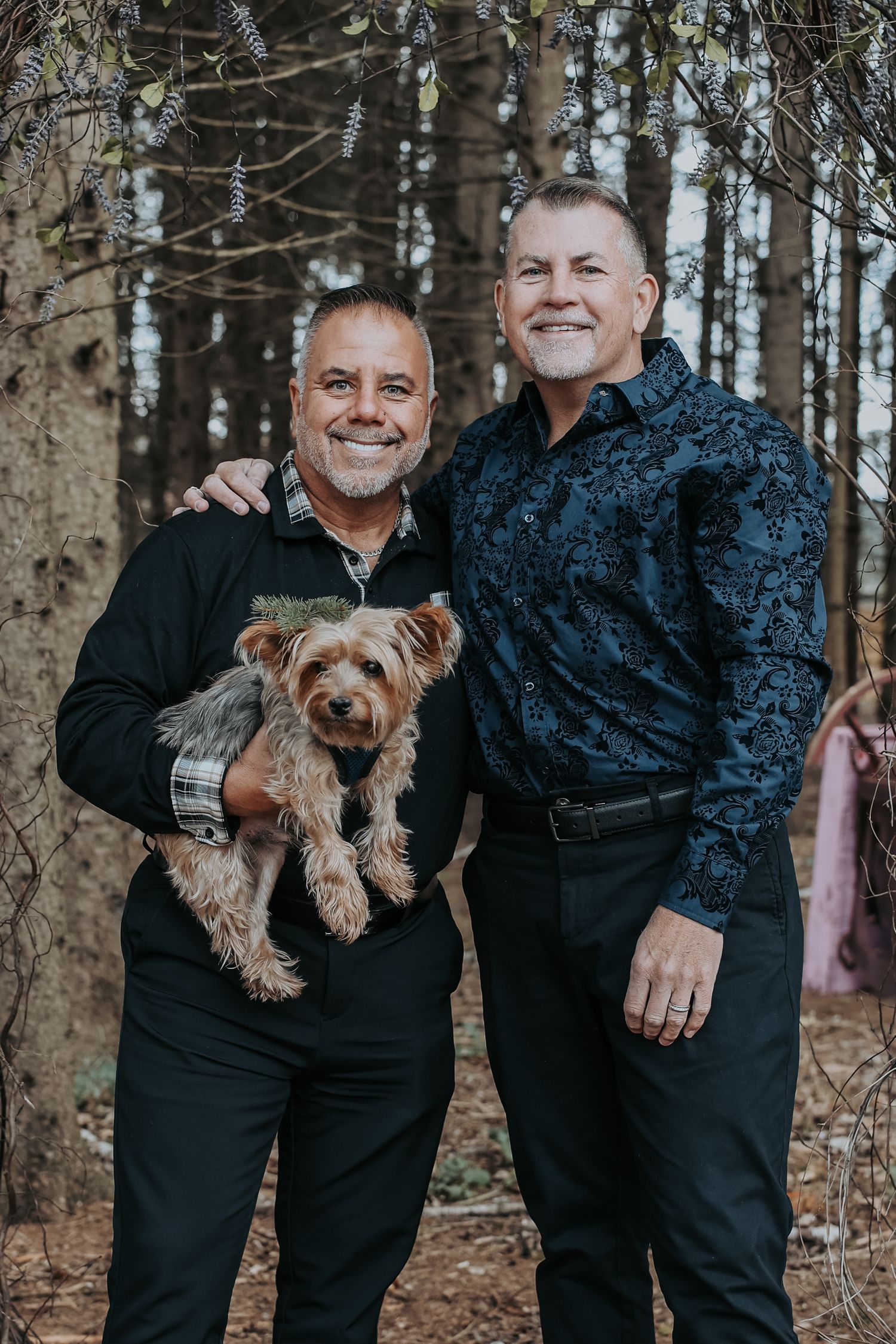 Couple in black outfits posing with their small brown dog in a wooded forest setting during autumn.