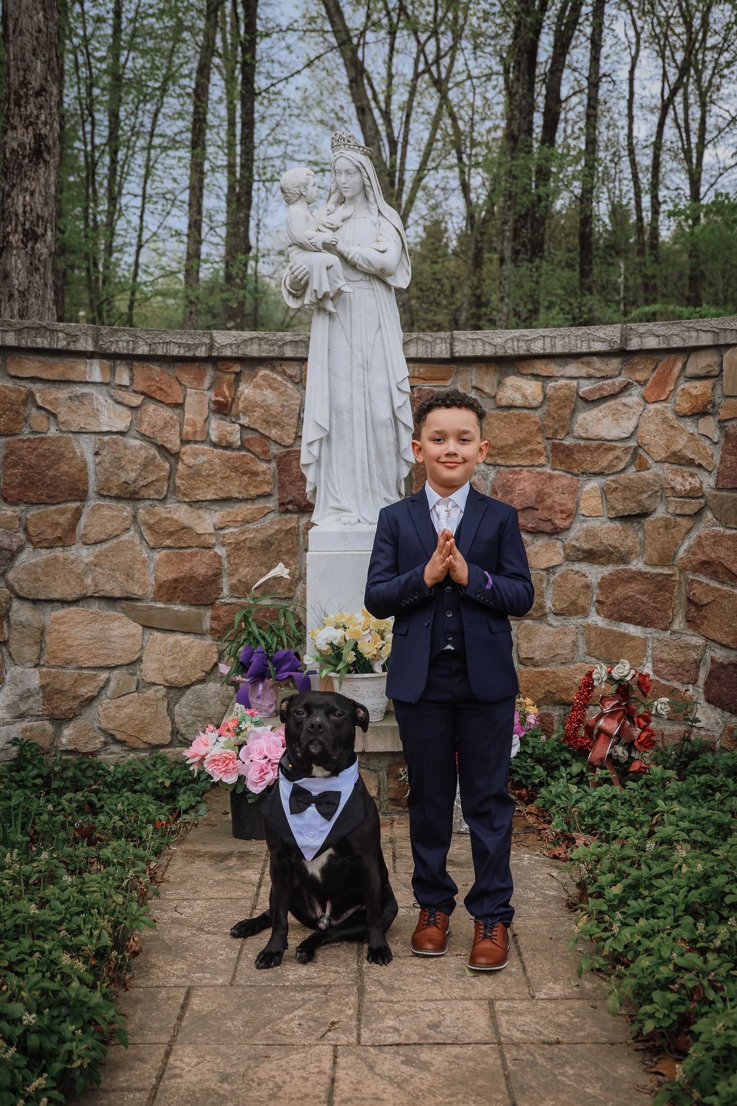 A child in a navy suit poses for First Communion with a black dog in front of a religious statue and stone wall.