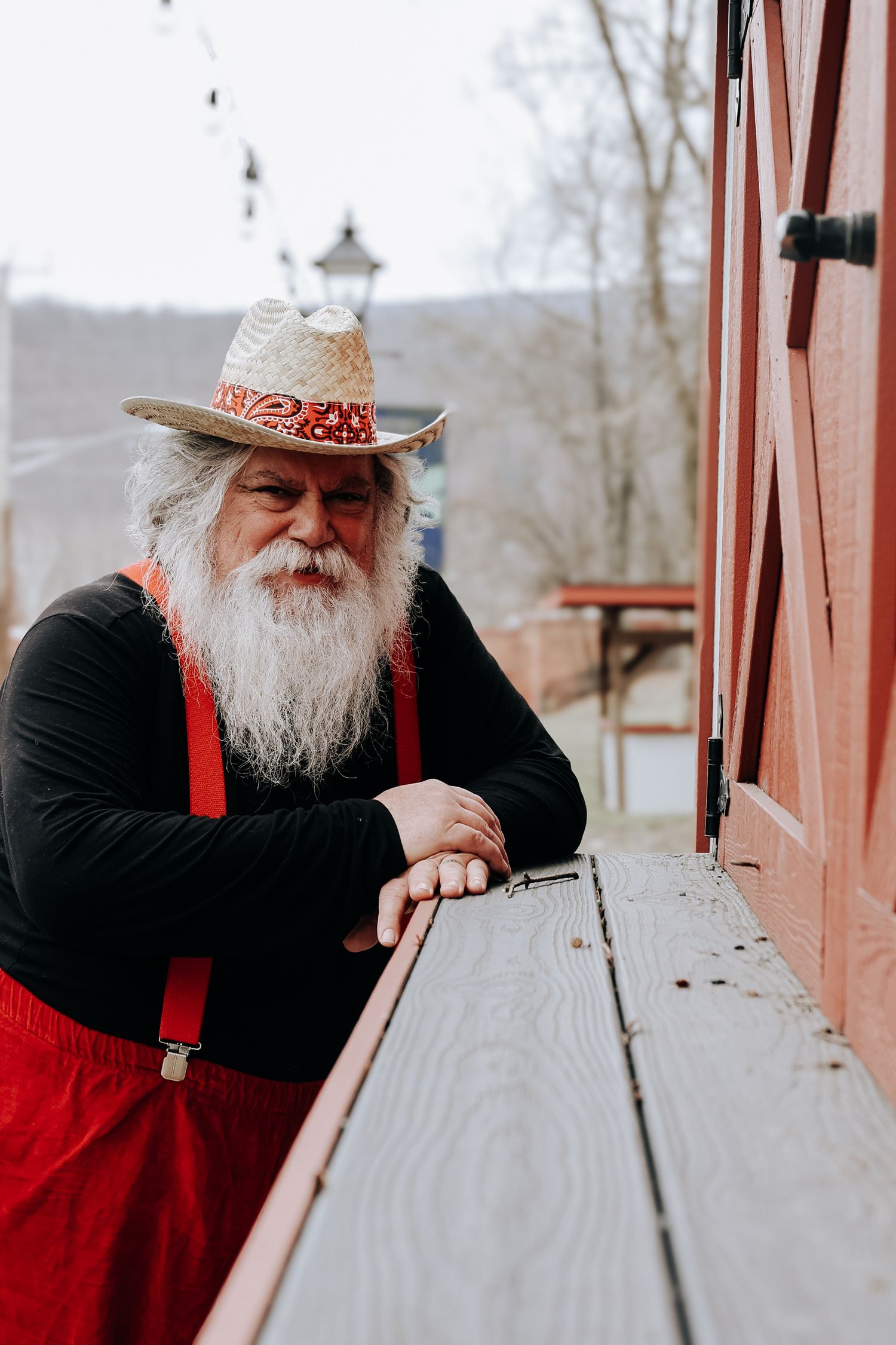 Person with a white beard wearing a cowboy hat and black shirt leans on a wooden railing outdoors.