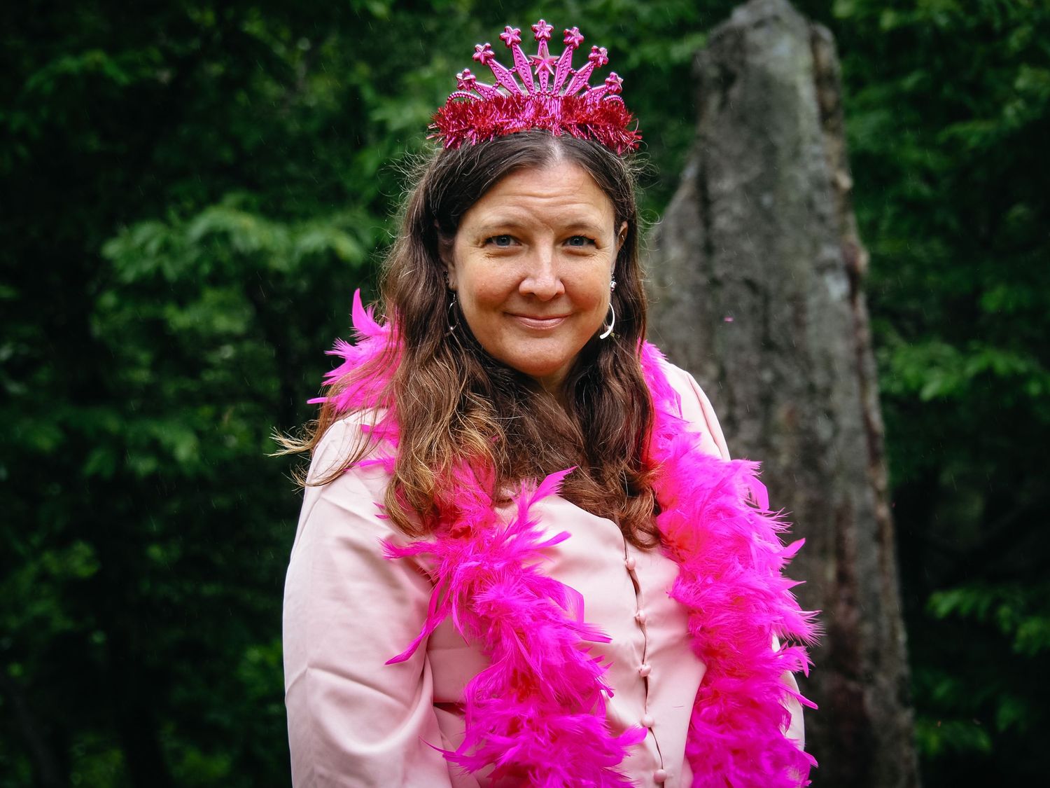 A person wearing a pink feather boa and tiara poses outdoors against a green forest backdrop.