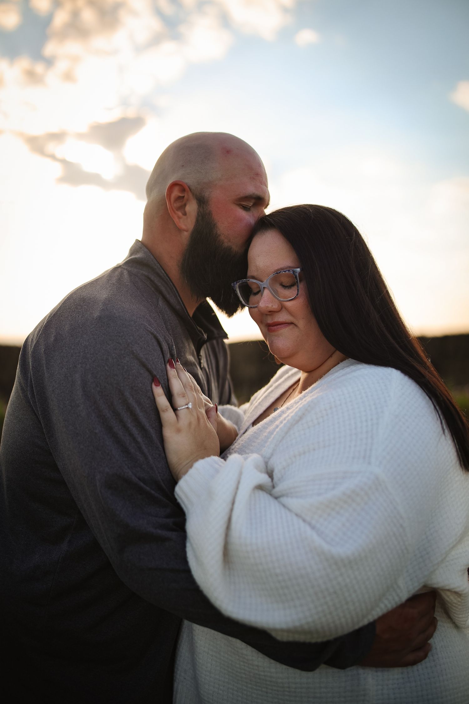 Intimate couple embraces at sunset, with woman in white sweater and man in gray shirt against soft sky backdrop.
