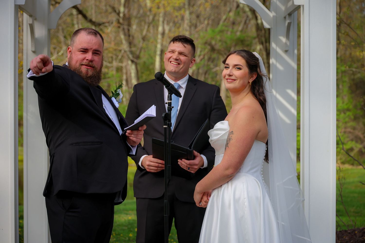 Bride and groom stand with officiant under white arbor during outdoor wedding ceremony in lush green garden.