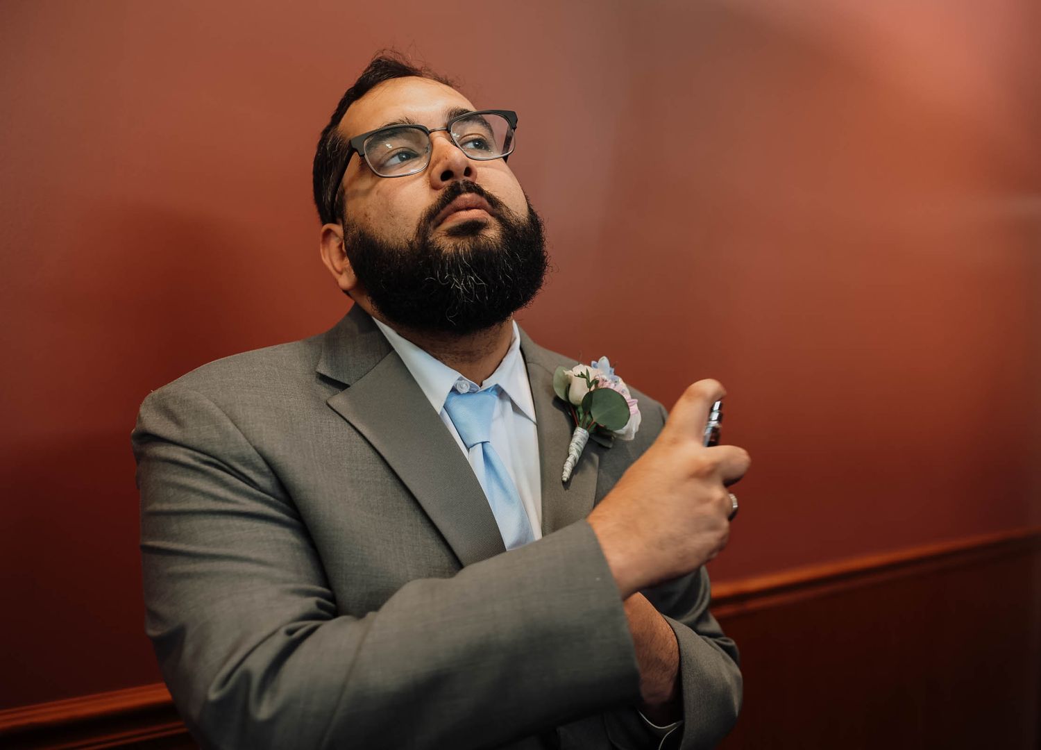 Bearded man in gray suit with boutonniere adjusting his tie before a formal event.