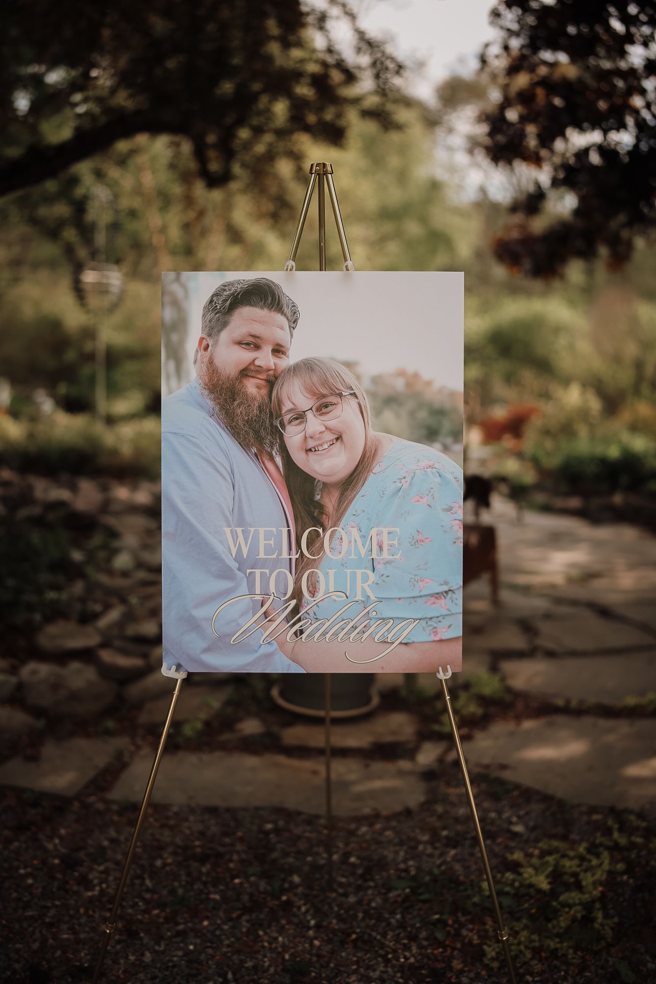 A wedding welcome sign on a gold easel displays a couple's photo outdoors, reading Welcome to Our Wedding.