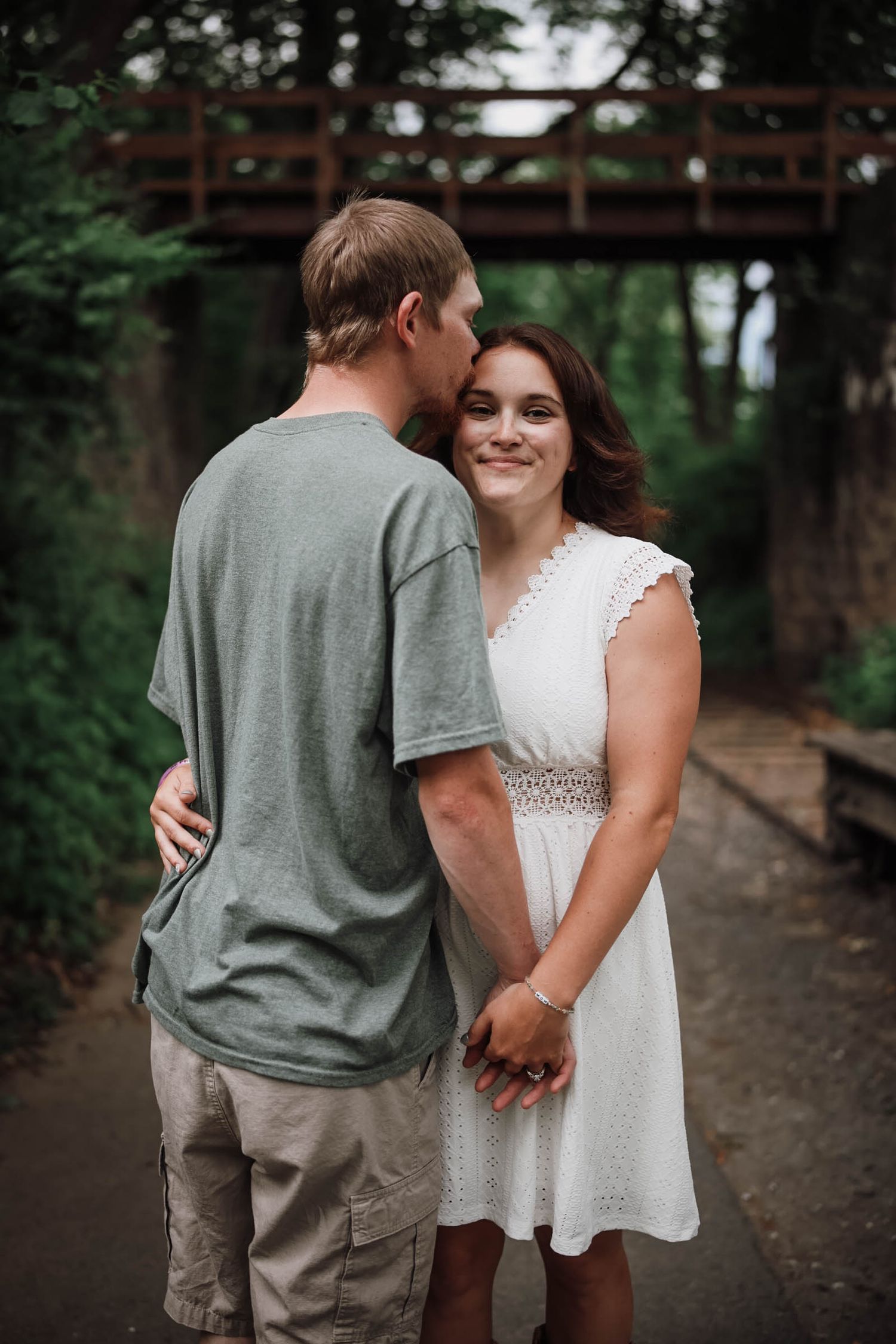 A couple shares a tender moment on a wooded path, man kissing woman's cheek as she smiles, wearing white dress.