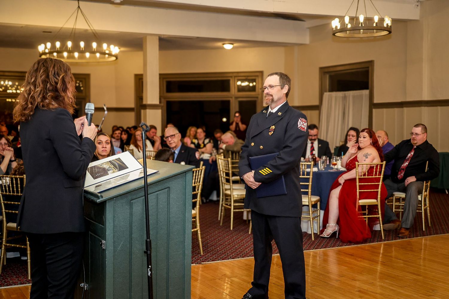 Fire chief receives award at formal banquet ceremony, standing before podium in dress uniform with guests seated behind.