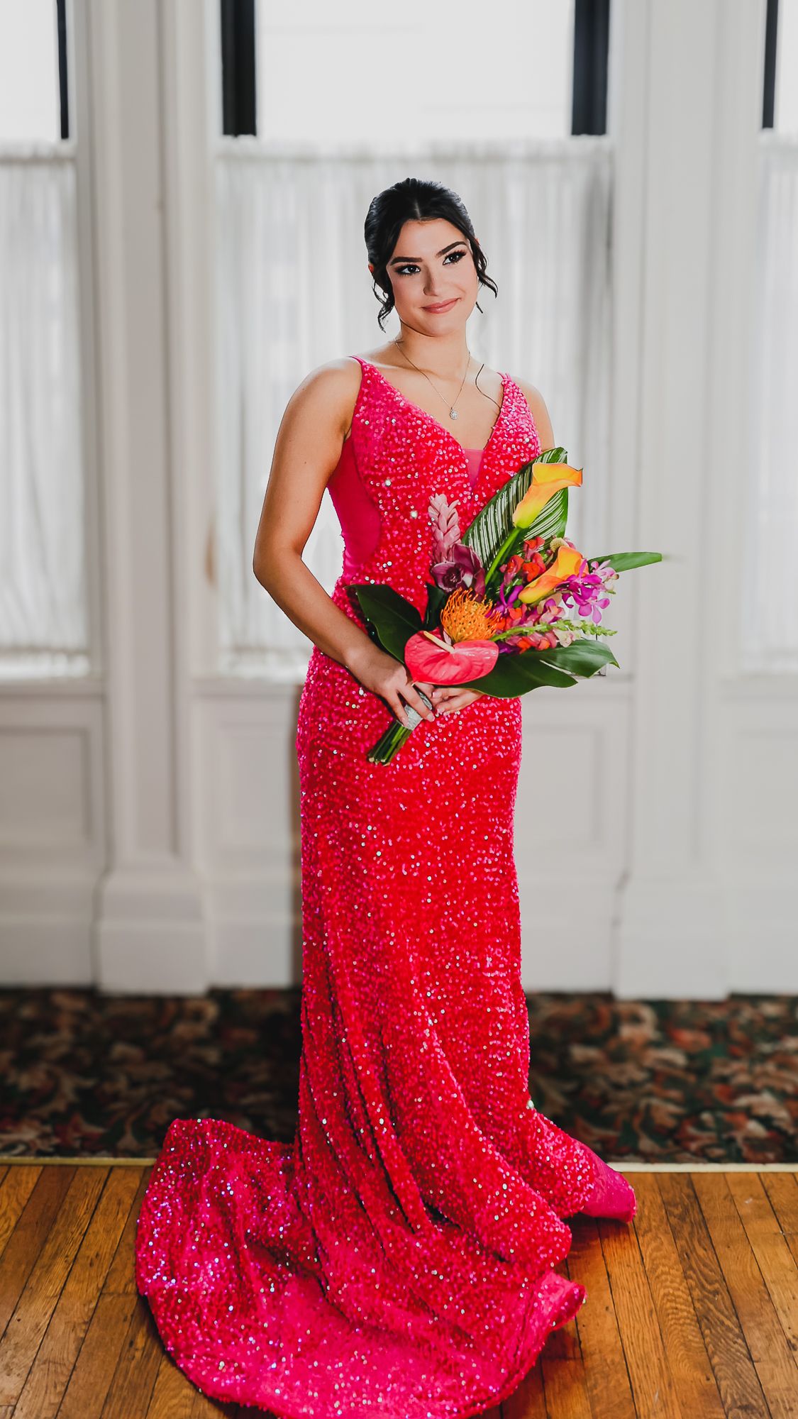 Woman in a hot pink sequin gown holding a tropical bouquet of colorful flowers, posing indoors.