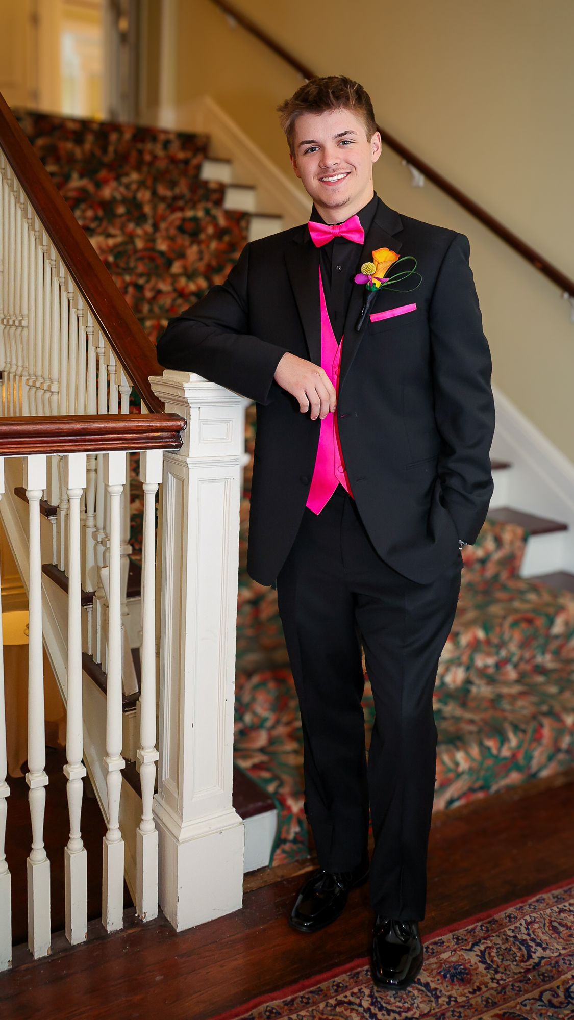 Young man in black tuxedo with hot pink bow tie, vest, and boutonniere posing by staircase railing.
