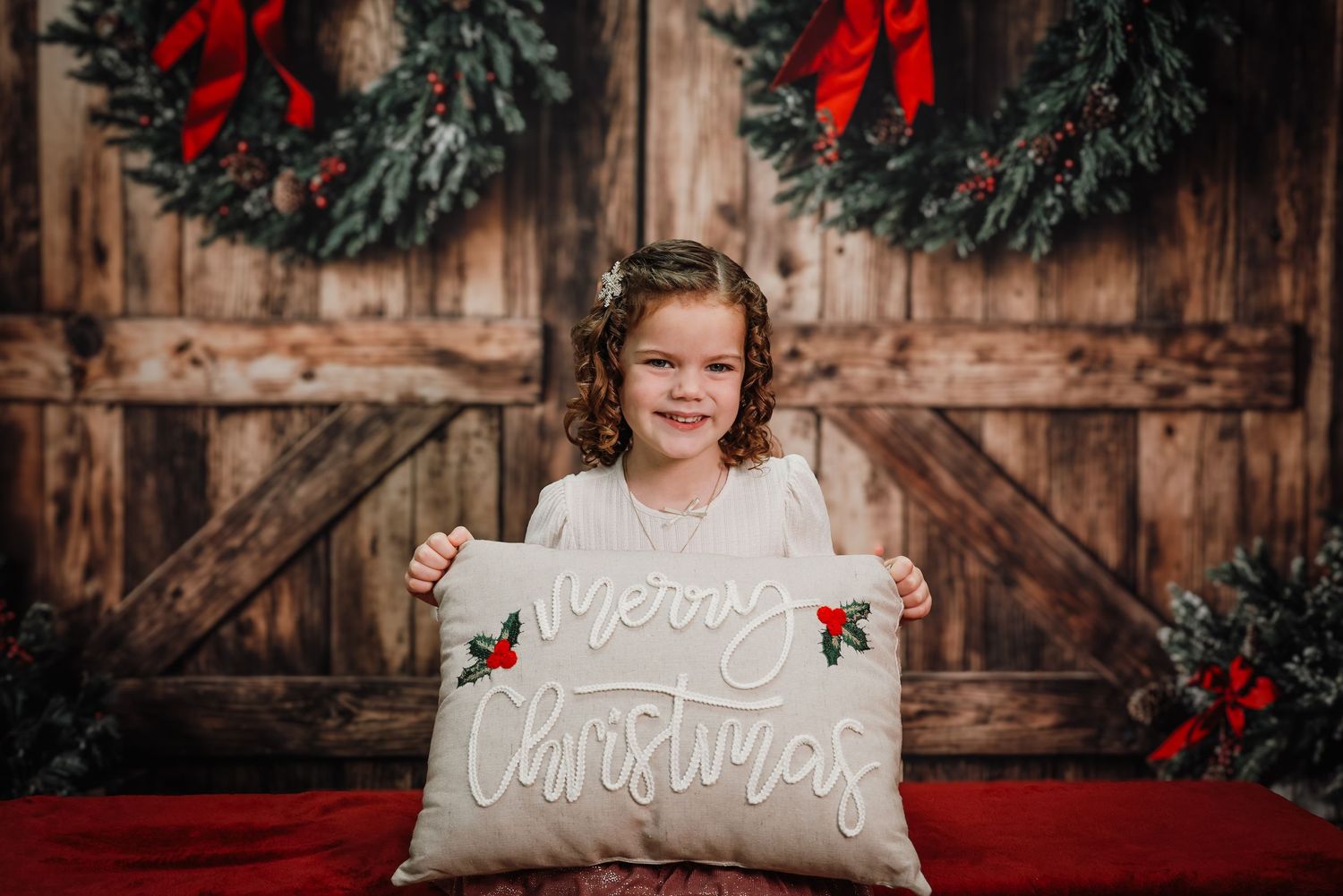 Young girl holding a Merry Christmas pillow in front of a rustic barn door backdrop with holiday wreaths.