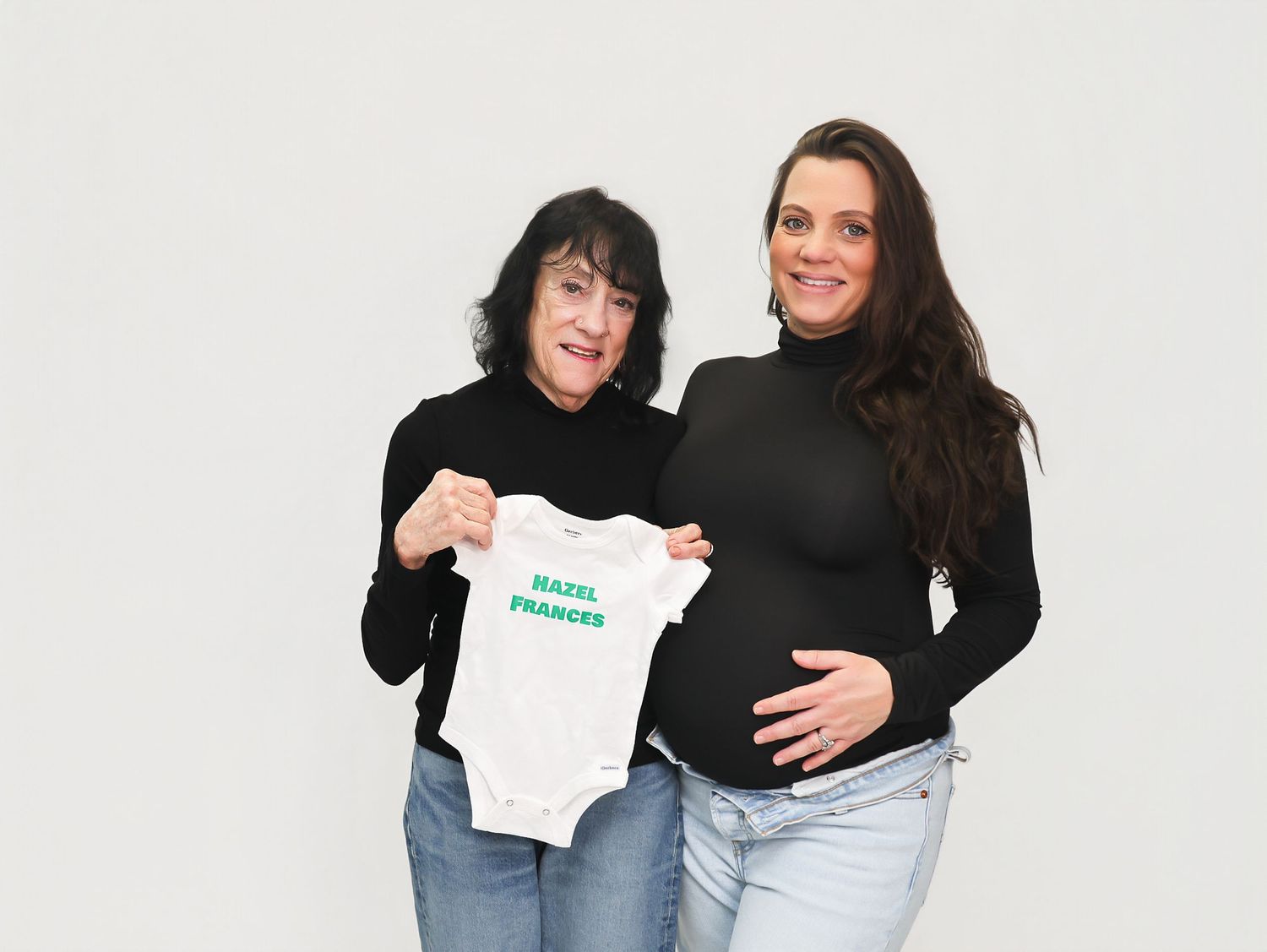 Two women pose together, one pregnant holding a white baby onesie reading Hazel Frances, both wearing black turtlenecks.