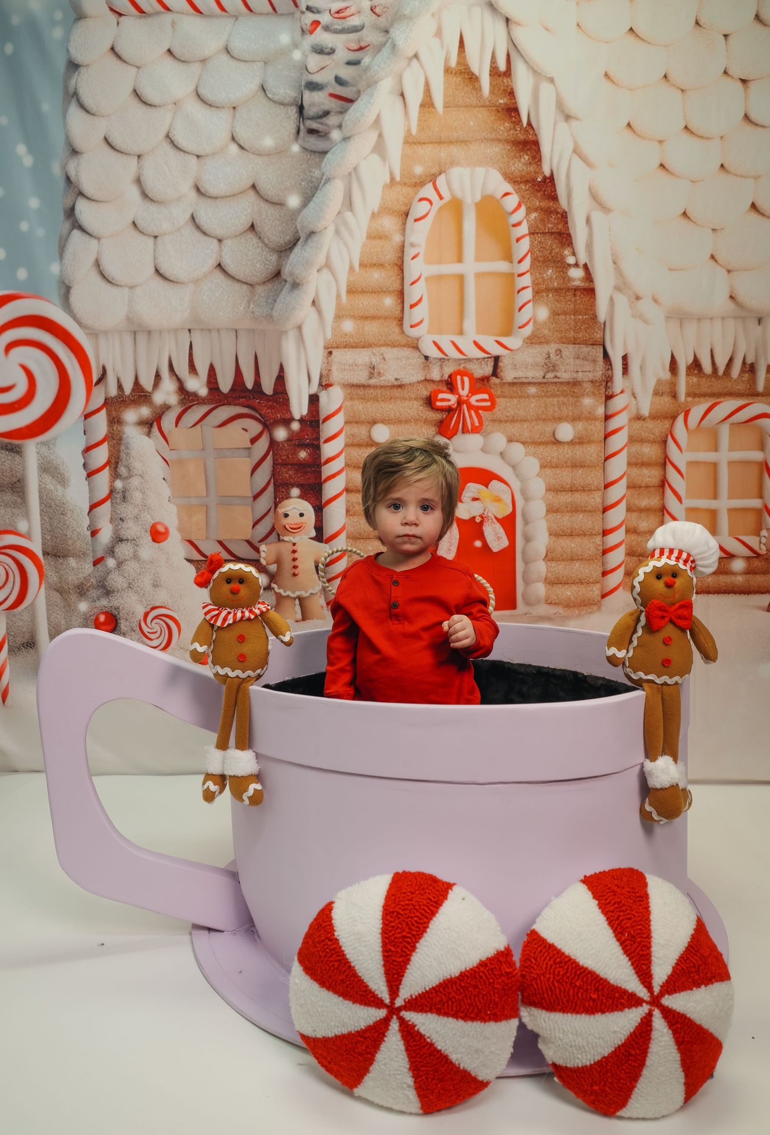 Toddler in red outfit sitting in oversized mug prop with gingerbread decor and candy house backdrop.
