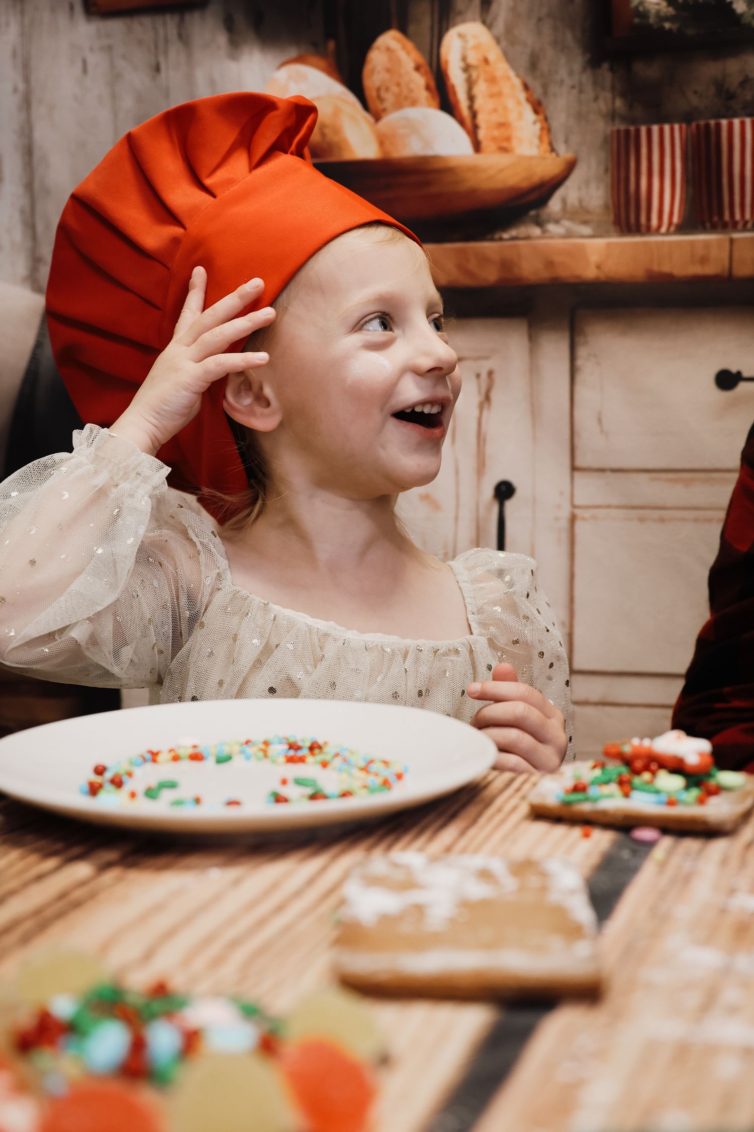 Excited young girl in red chef hat decorating cookies with colorful sprinkles in a rustic kitchen.