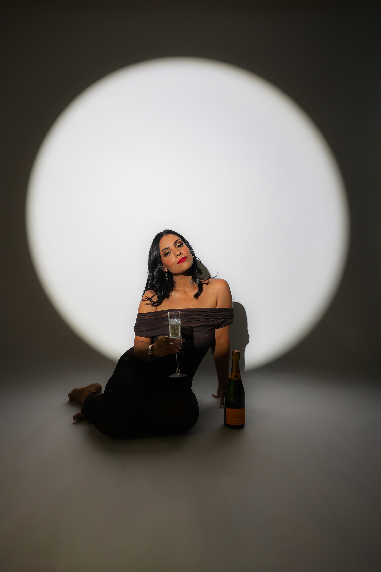 Woman in off-shoulder dress sits on floor holding champagne glass with bottle nearby, posed before circular light backdrop.