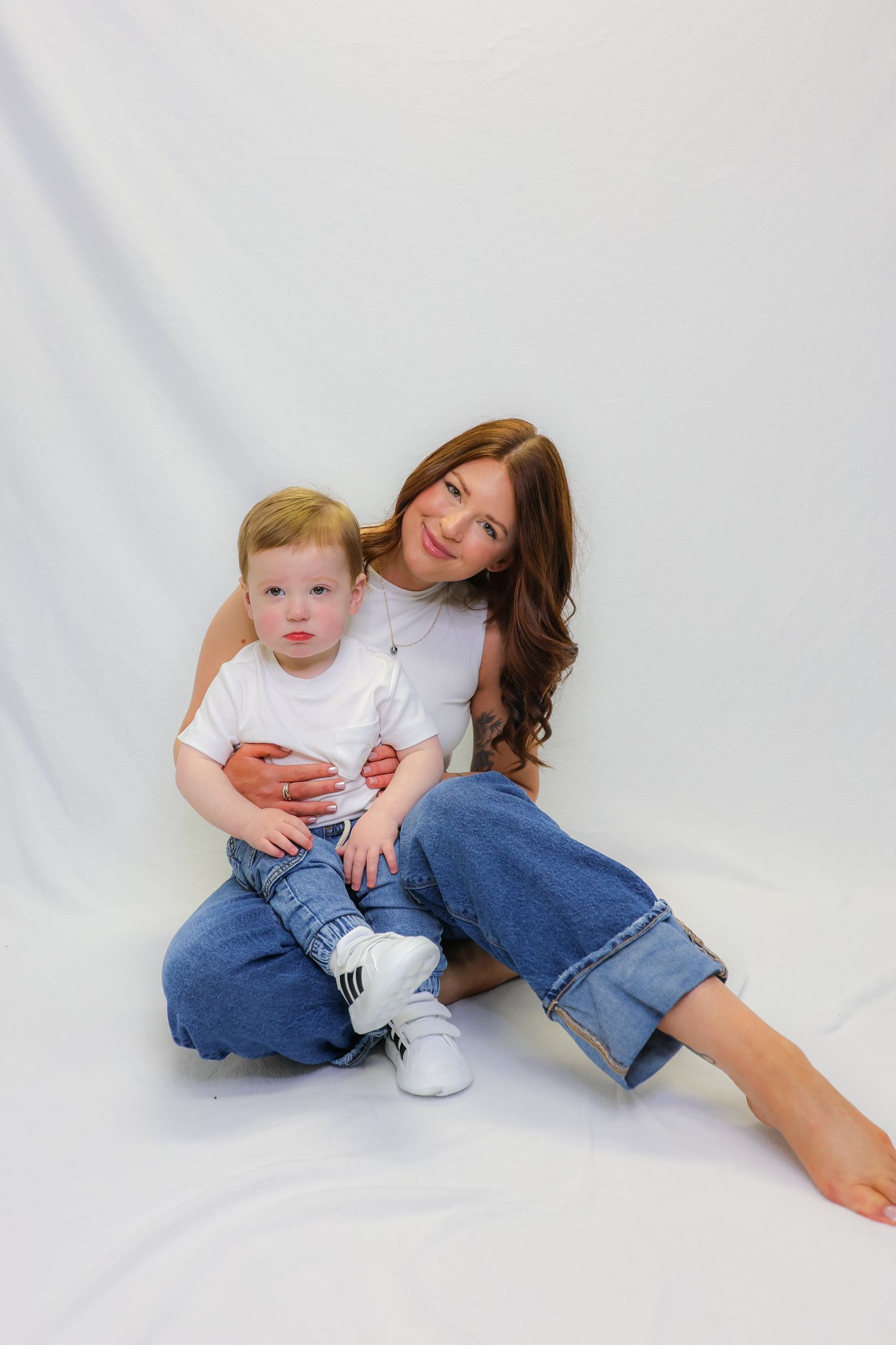 A young woman with long brown hair sits on white backdrop hugging a toddler, both wearing white tees and jeans.