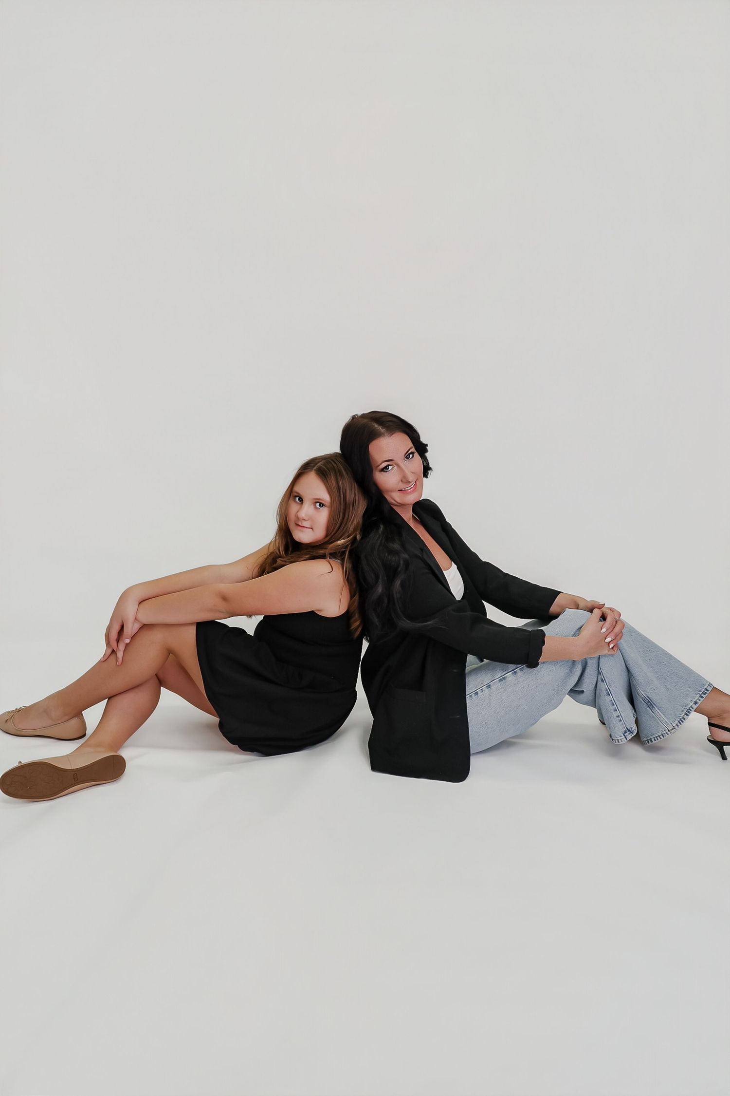 Two women in black outfits sit back-to-back on a white studio floor, posing confidently for a fashion photo.