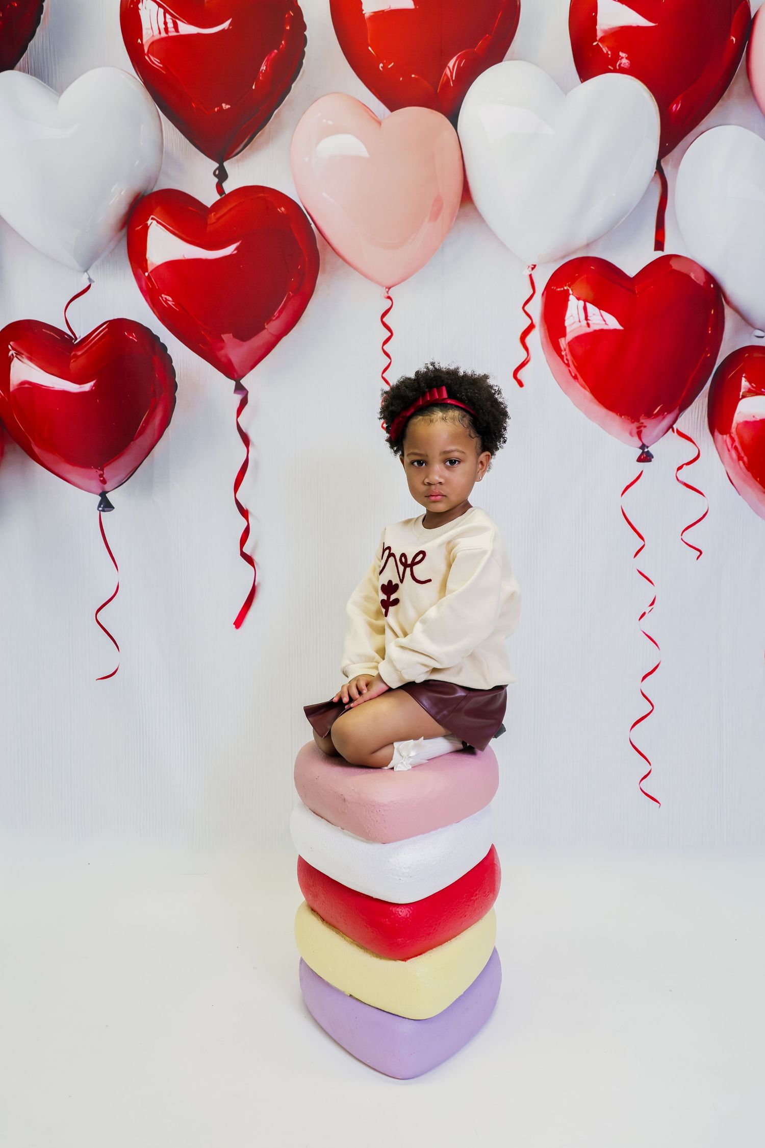 A young girl sits atop stacked heart-shaped boxes surrounded by red, pink, and white heart balloons in a Valentine's Day photo.