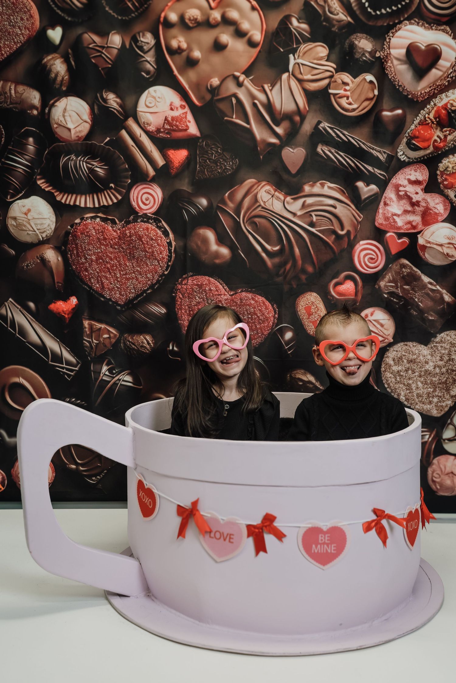 Two children wearing heart-shaped glasses sit in a giant teacup prop with Valentine's Day chocolate backdrop.