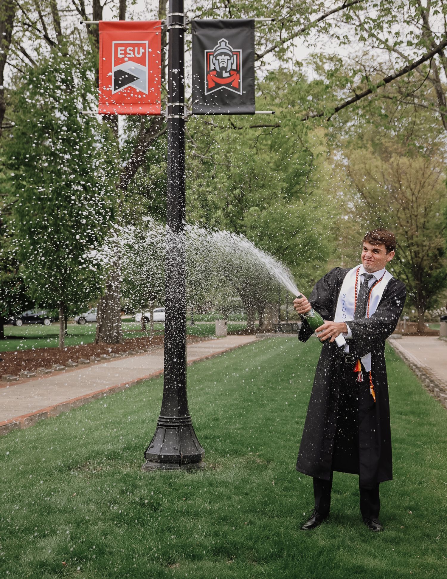Graduate in academic regalia pops champagne bottle outdoors on college campus with ESU banners on lamppost.