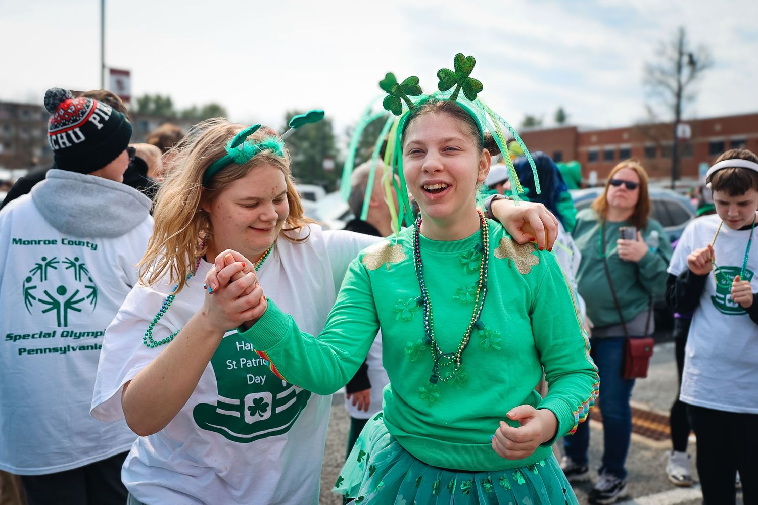 Two women in green St. Patrick's Day outfits smile and dance at an outdoor Special Olympics parade event.