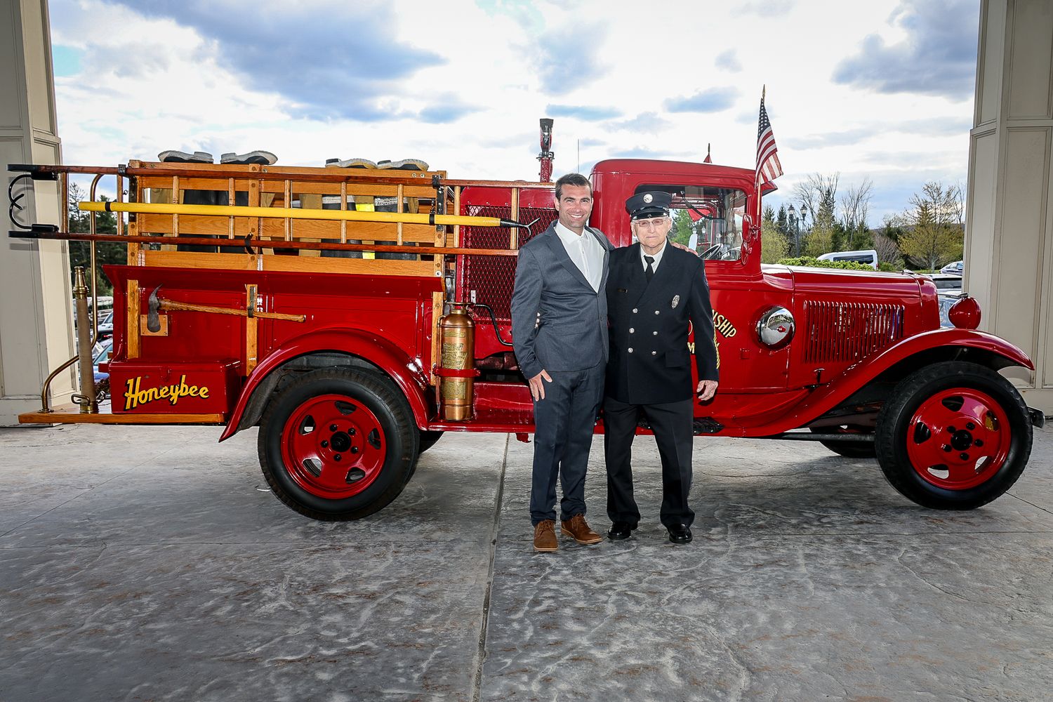 Two men posing in front of a vintage red Hampden fire truck displayed outside a fire station.