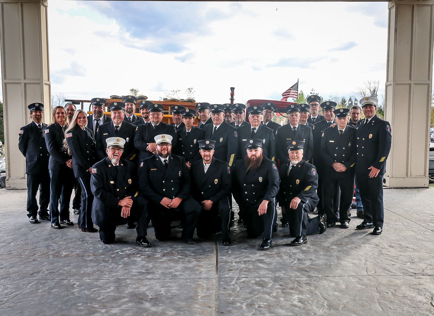 Group of firefighters in dress uniforms posing for a formal photo outside a fire station with a fire truck visible behind them.