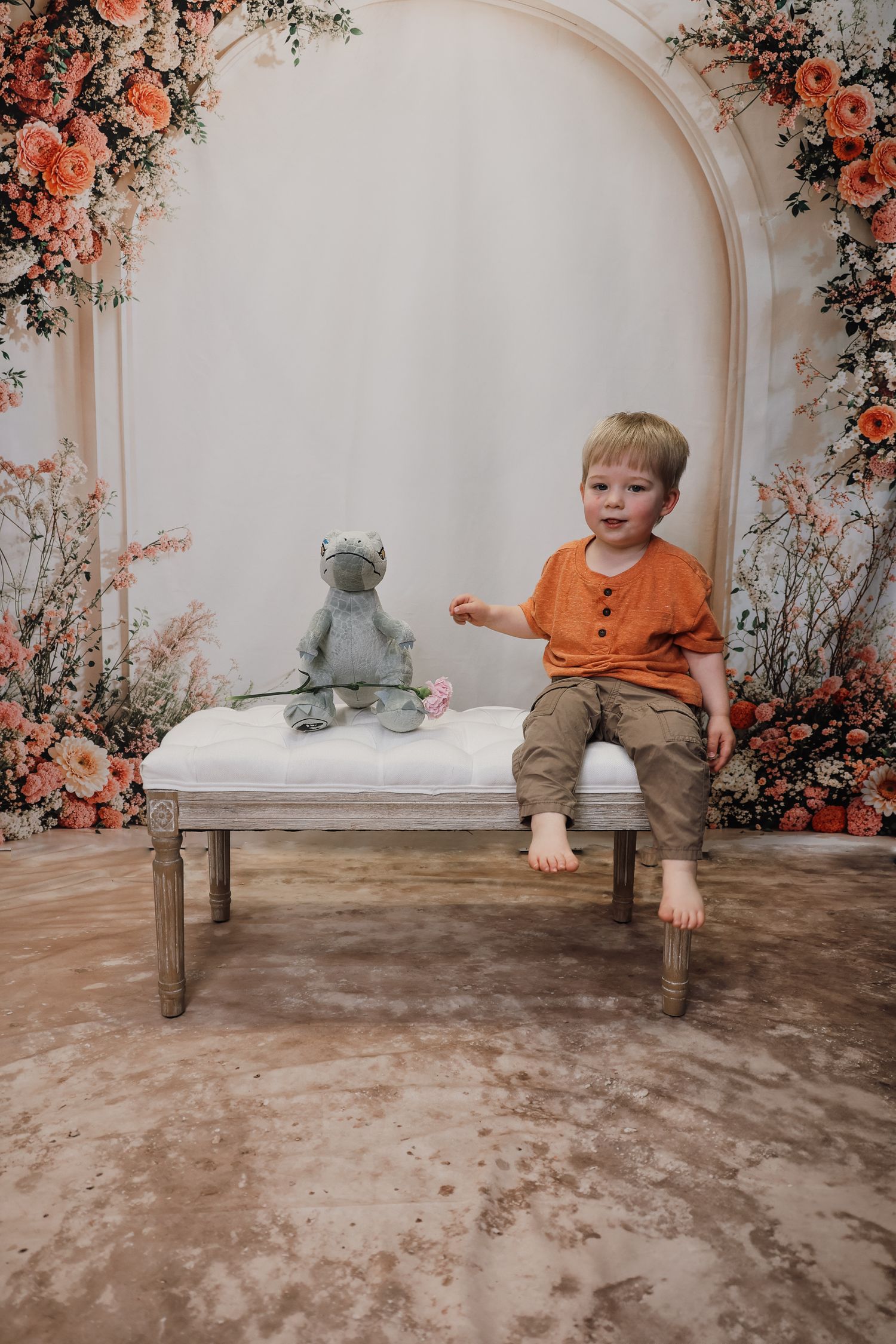 Young boy in orange shirt sits on white bench with stuffed animal toy, surrounded by floral arch backdrop.