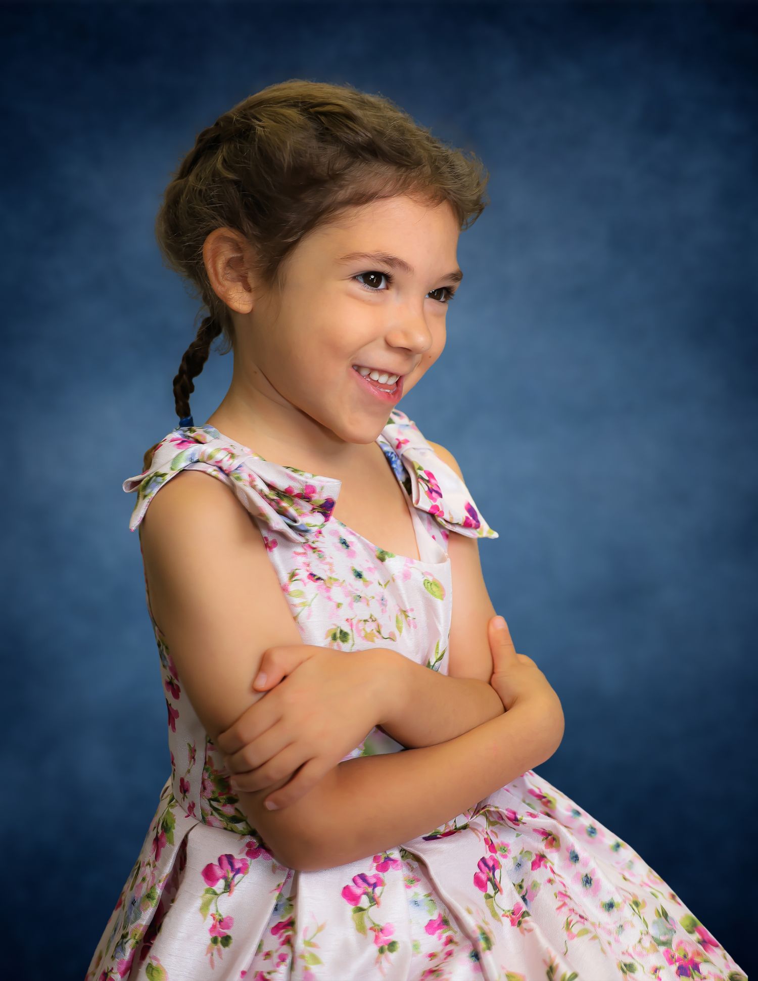 Young girl in floral dress with braids poses confidently with arms crossed against blue studio background.