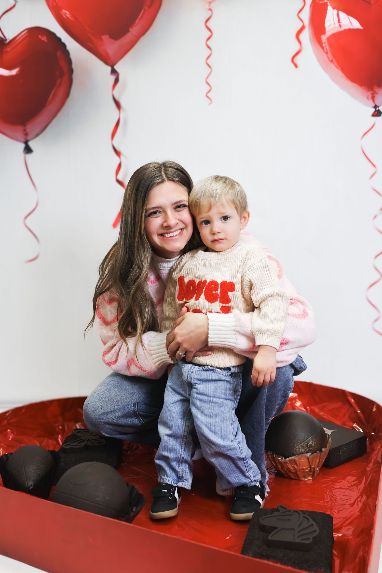 Young woman and toddler boy sit in red heart box with chocolate, surrounded by red heart balloons for Valentine's Day.