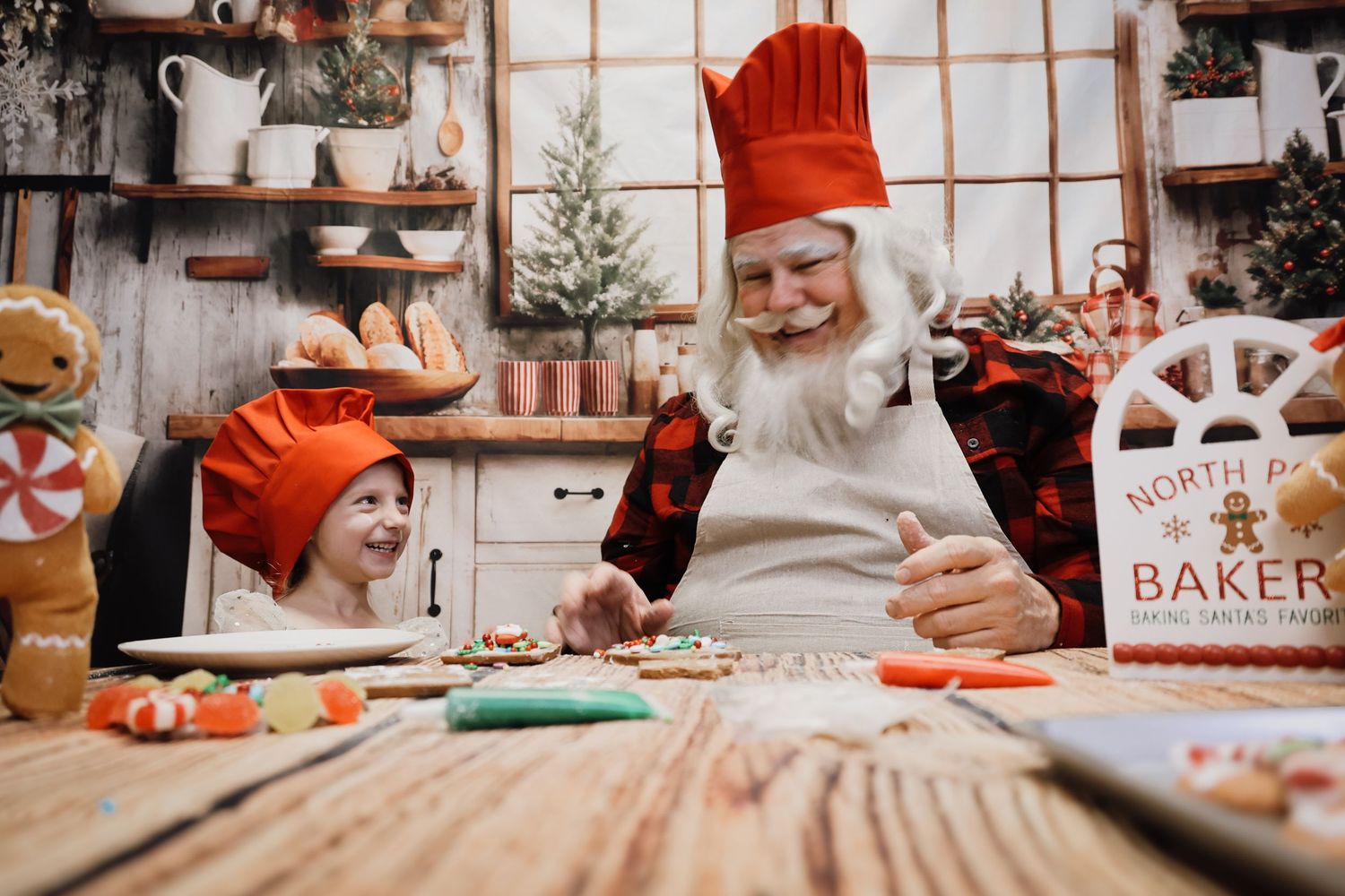 Santa Claus and child in chef hats decorating Christmas cookies together in a festive holiday bakery kitchen.
