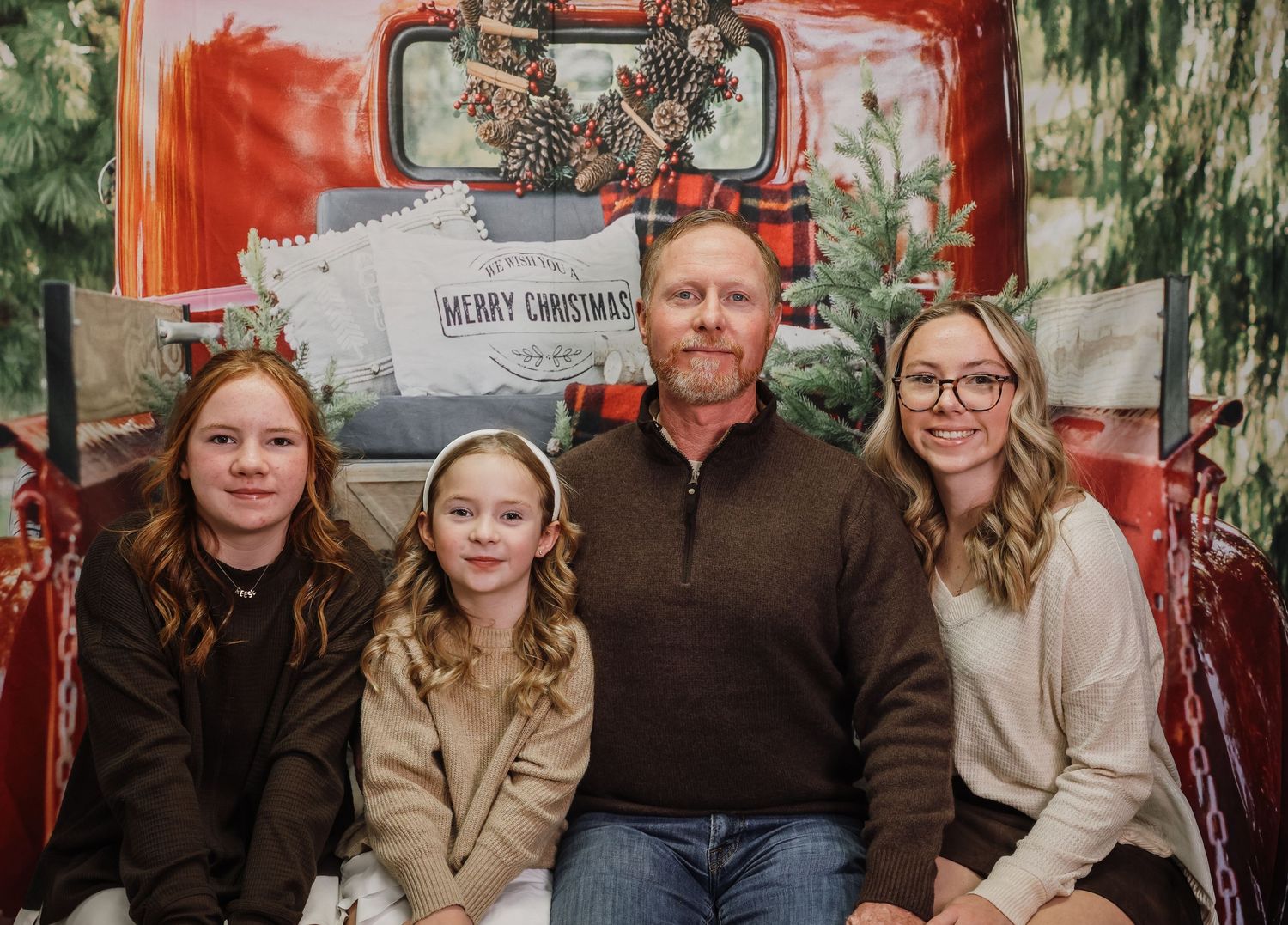 Family of four posing for a Christmas photo in front of a festive red truck backdrop with holiday decorations.