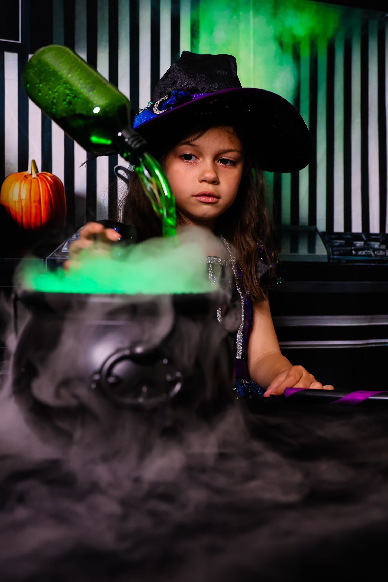Young girl in witch hat pouring green potion into smoking cauldron for Halloween costume celebration.