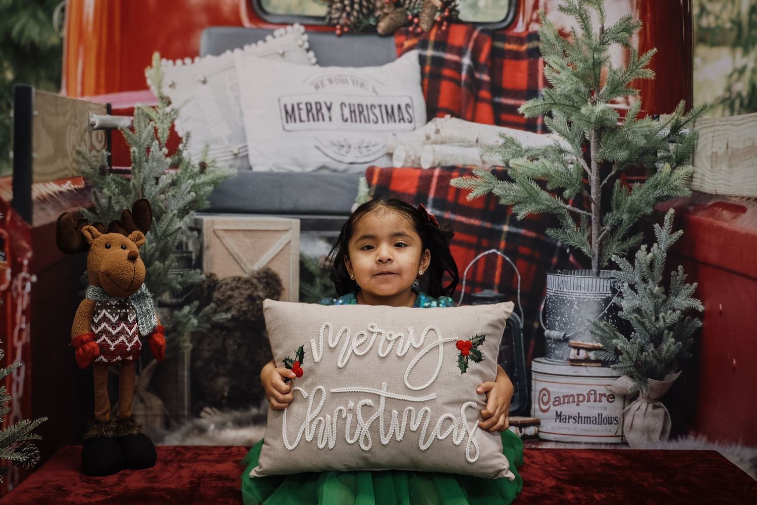 Young girl holding a Merry Christmas pillow in front of a festive holiday backdrop with a red truck and Christmas trees.