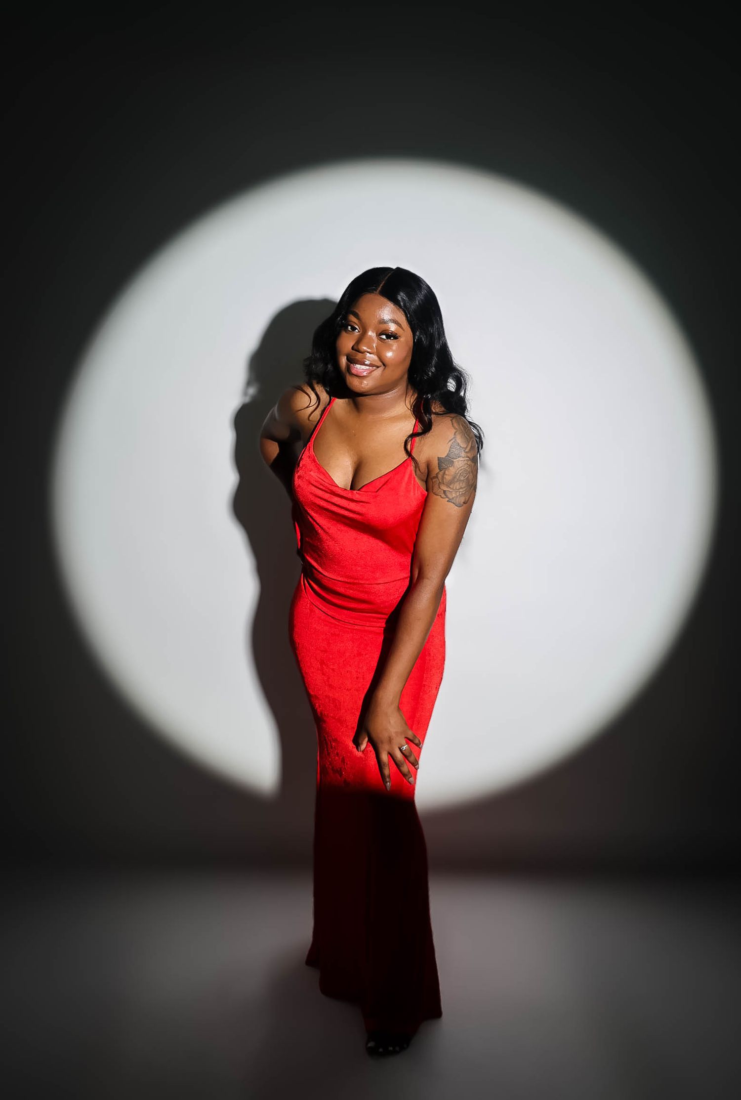 A woman in a red gown poses confidently in a spotlight against a dark background, showcasing a tattoo on her arm.