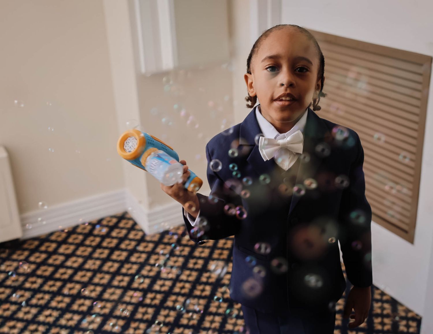Young boy in navy suit and bow tie blowing bubbles with a bubble gun indoors at a formal event.