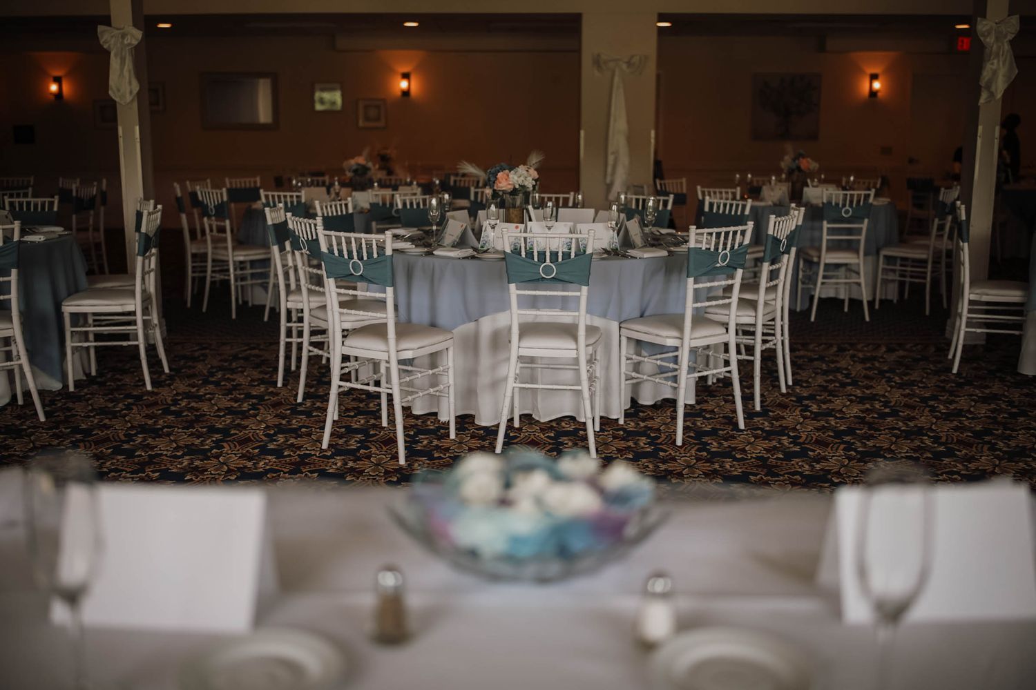 Elegant banquet hall with round tables draped in blue linens, white chiavari chairs, and floral centerpieces for a wedding reception.