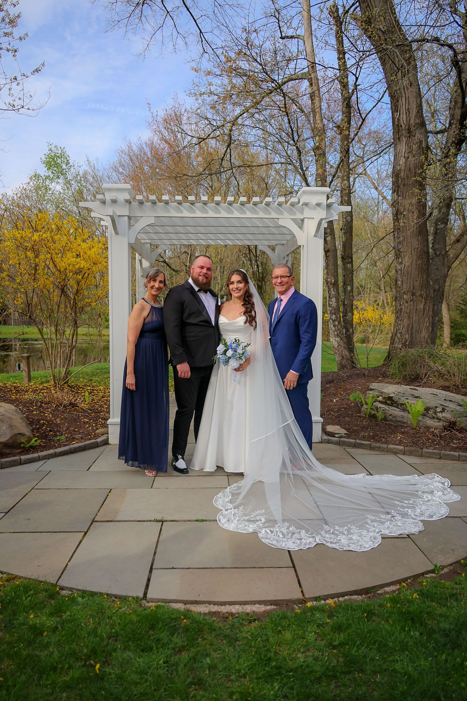 Bride in white gown with veil poses with groom and family under white pergola in garden setting.
