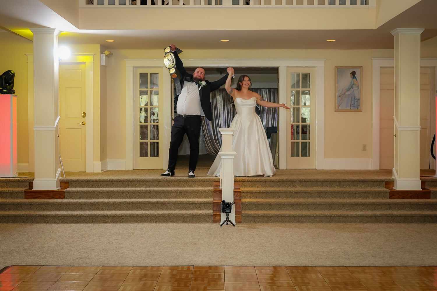 Bride and groom joyfully dancing at wedding reception, raising hands in celebration near elegant staircase.