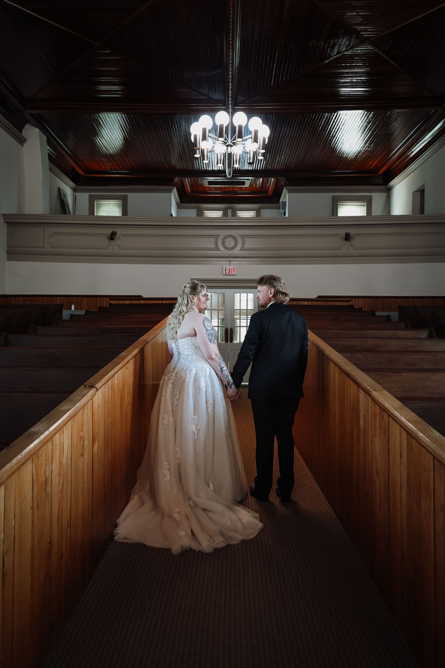 Bride and groom hold hands walking down church aisle, chandelier above, wooden pews on both sides.