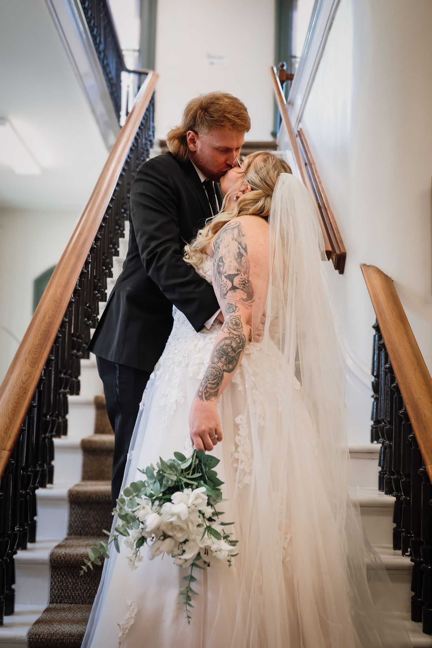 Bride and groom share a kiss on elegant staircase, bride wearing lace gown with veil holding white floral bouquet.