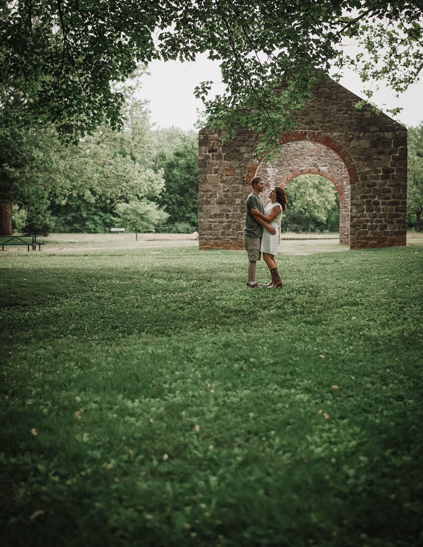 Couple embracing in lush green park beside rustic brick archway ruins, surrounded by trees.