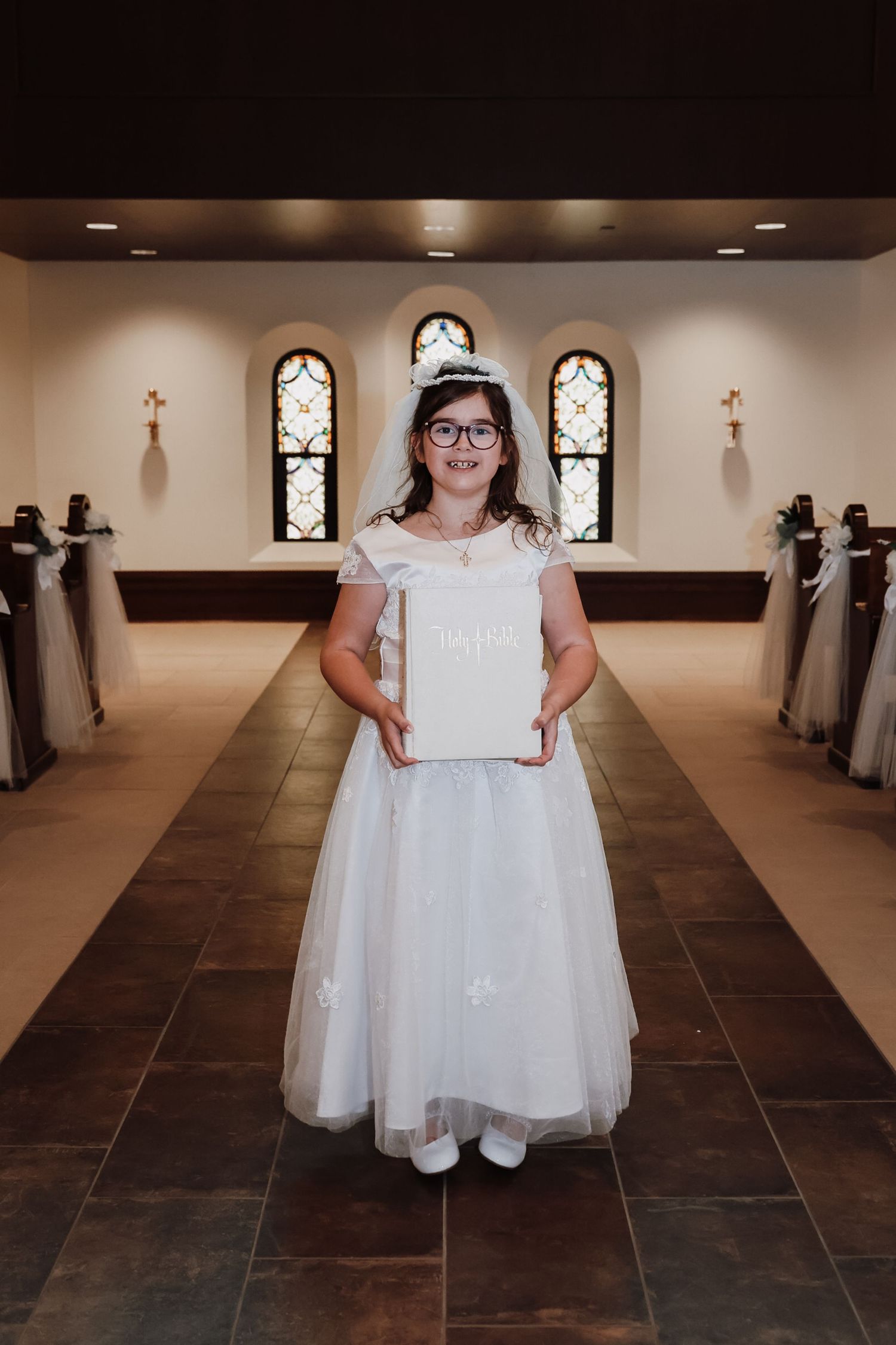 Young girl in white dress and tiara holds a Bible in a church aisle during her First Communion ceremony.