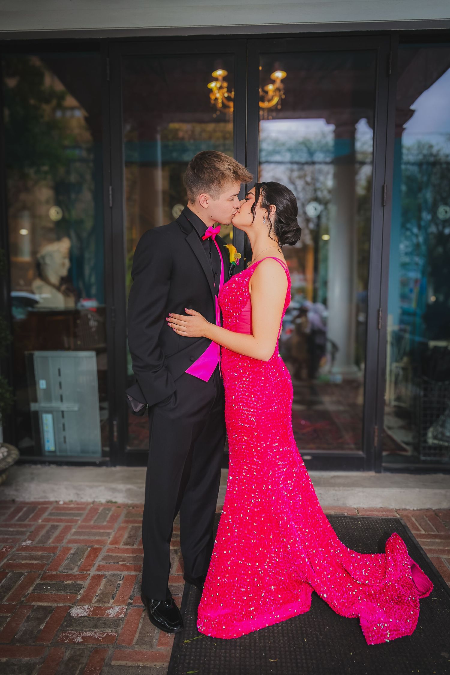 Couple sharing a kiss at prom, woman in red sequin gown and man in black tuxedo with pink bow tie.