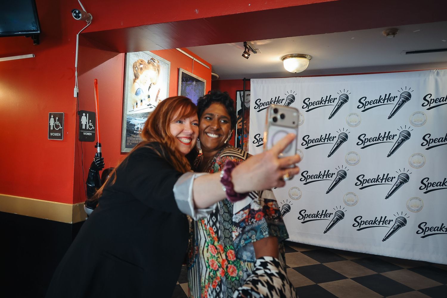 Two women smile and take a selfie together at a SpeakHer event in front of a branded step-and-repeat backdrop.