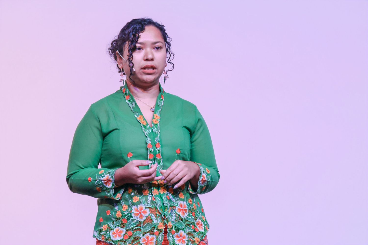 Woman in green floral cardigan presenting on stage against a soft purple background.