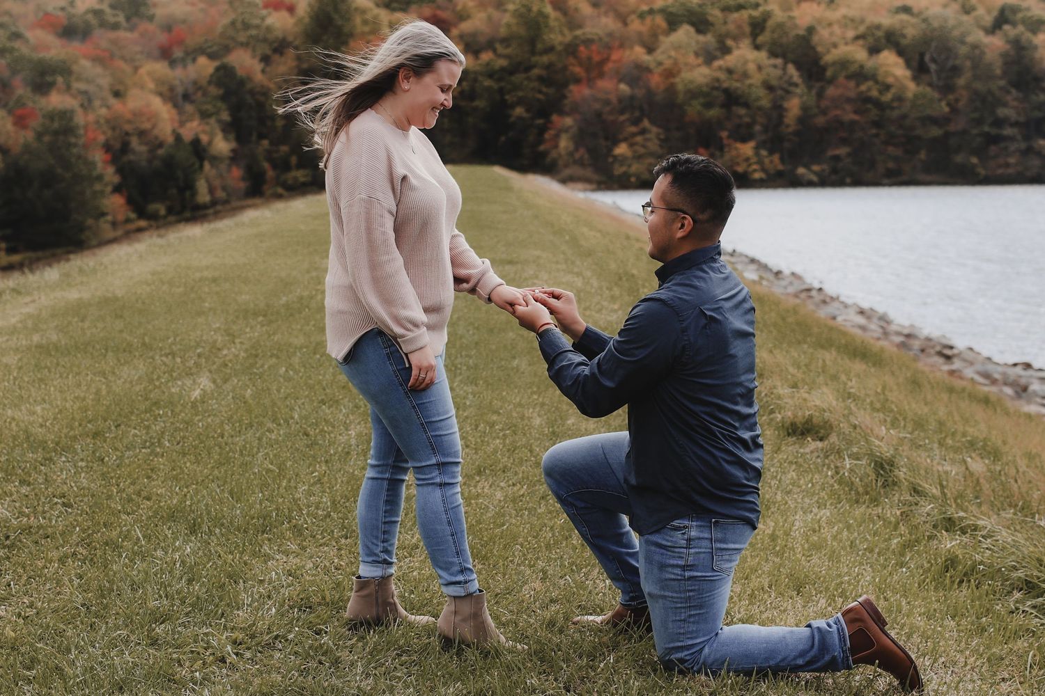 A romantic proposal moment captured on a grassy hillside during autumn with colorful fall foliage in the background.