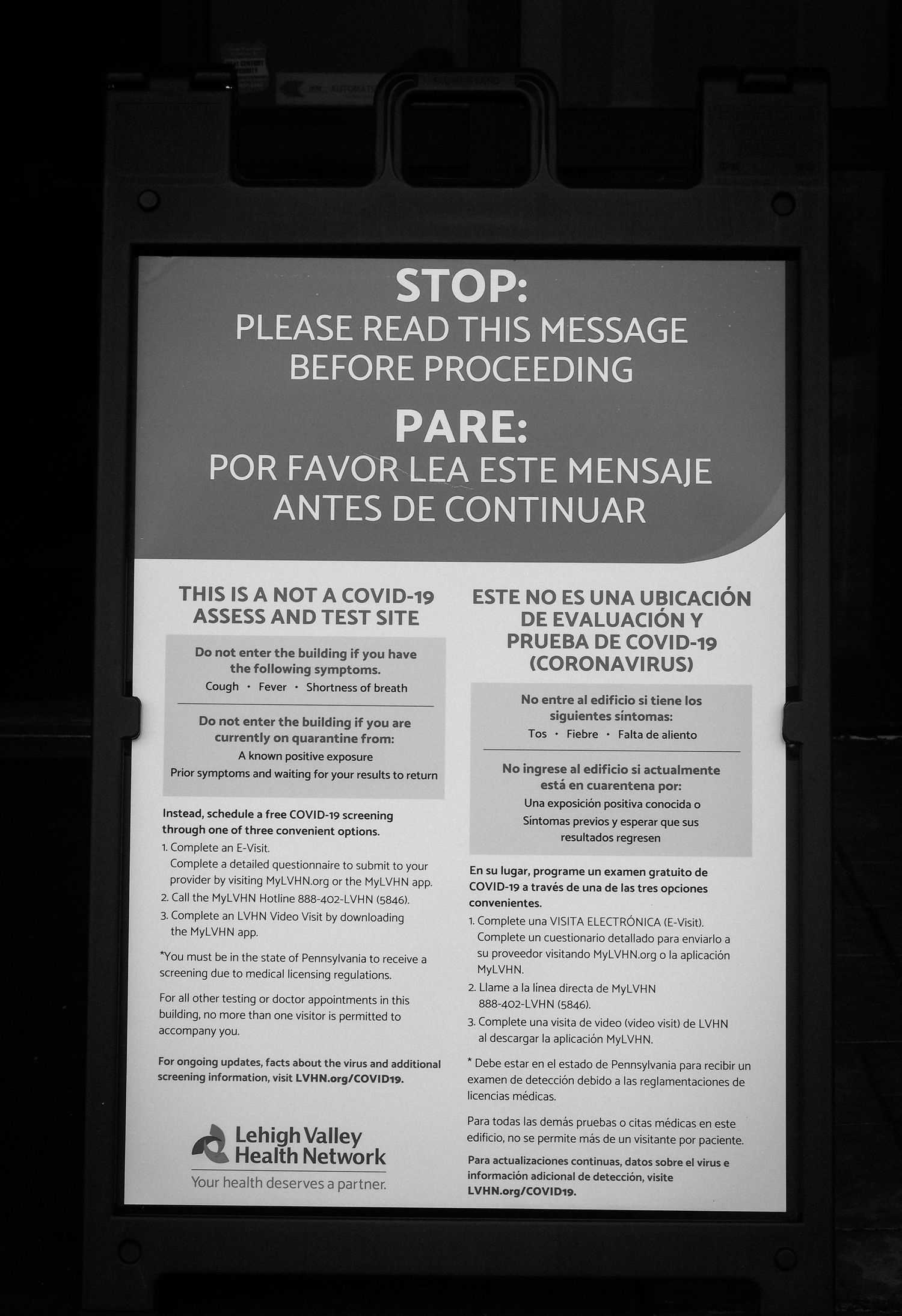 Black and white photo of a bilingual hospital sign with COVID-19 related instructions in English and Spanish.