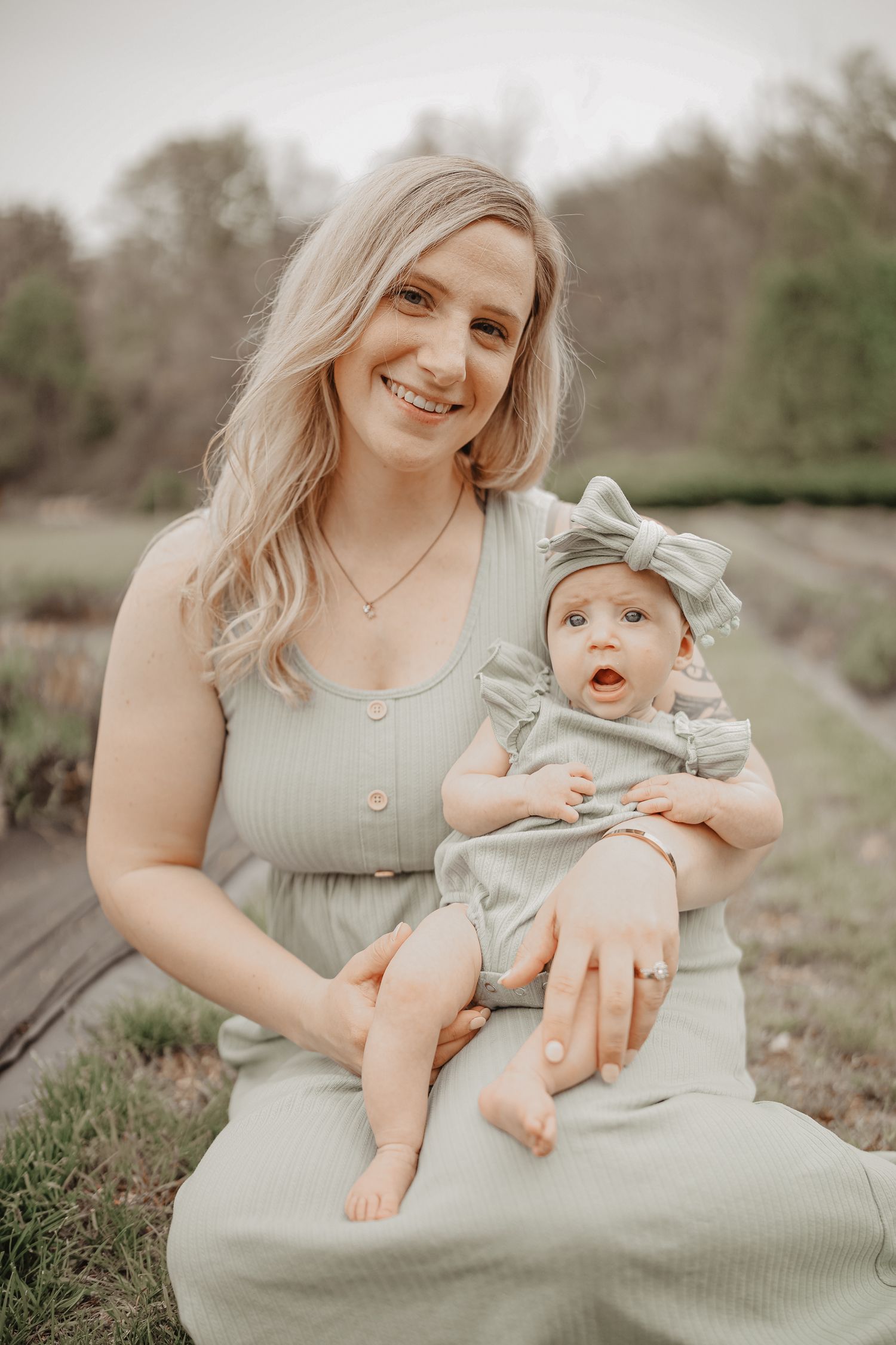 A parent wearing a gray dress sits in a grassy field holding a baby in matching gray outfit with a bow headband.