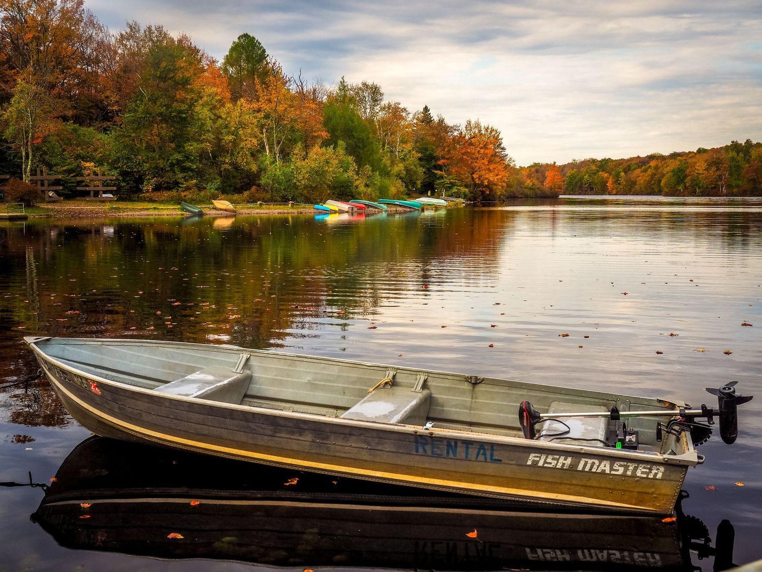 An aluminum fishing boat rests on a calm lake surrounded by vibrant autumn foliage and colorful kayaks on the shore.